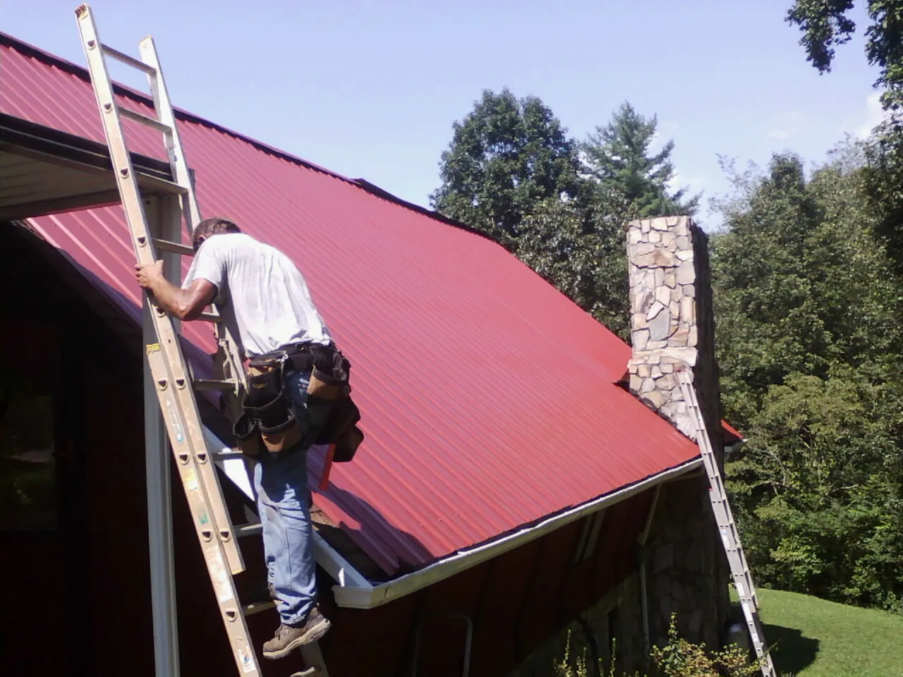 Roofer on a ladder beside a red metal roof and stone chimney — Soffit & Fascia Repair by Roofing Brothers in Croton Falls